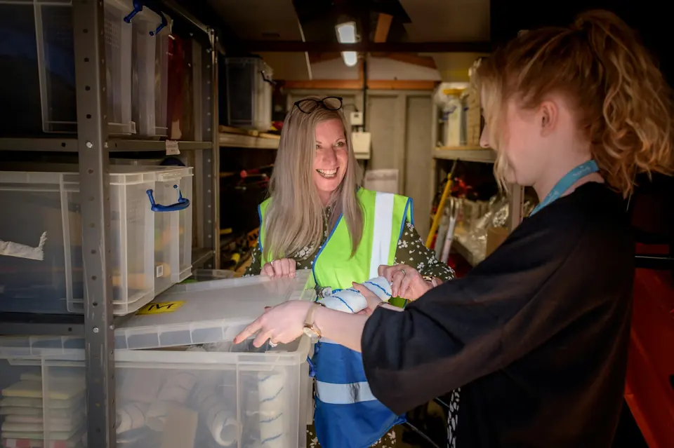 Smilende kvinne i refleksvest hjelper en kollega med å sortere plastkasser på et lager