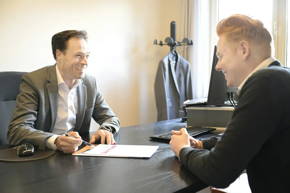 To smilende menn i samtale ved et skrivebord med et dokument mellom seg