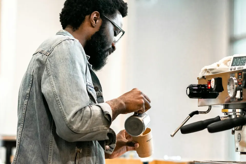 Mann i dongerijakke lager kaffe ved en espressomaskin på en lys kafé